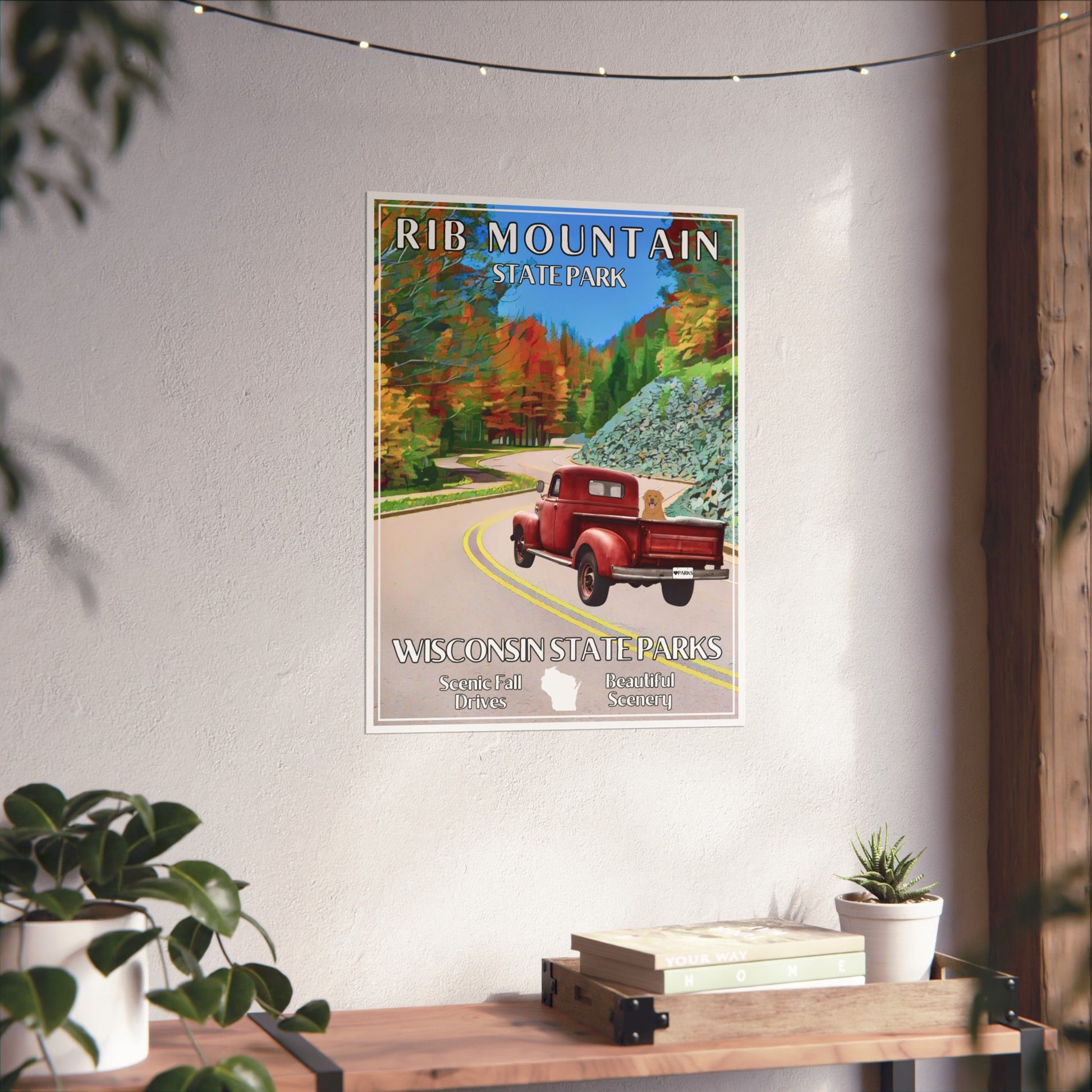 18x24” poster depicting a vintage red truck with a dog in the back driving up a curvy road lined with trees in vibrant color. Photo background: Poster hung on a wall above a shelf with books and plants.