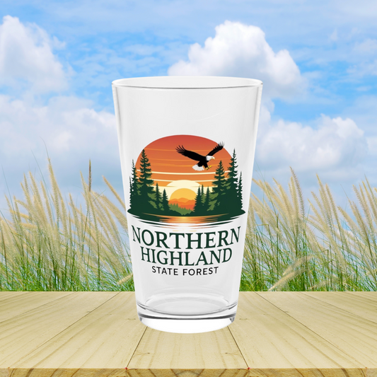 16 oz glass pint featuring a colorful design of a sunset over a lake surrounded by pine trees and a Bald Eagle Soaring overhead, with the text Northern Highland State Forest.  Photo background: glass sitting on wood boards in front of tall grasses.