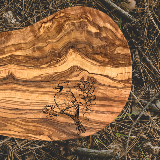 Olive wood serving board with an engraving of a cardinal sitting on a flowering branch.  Photo background: pine needles.