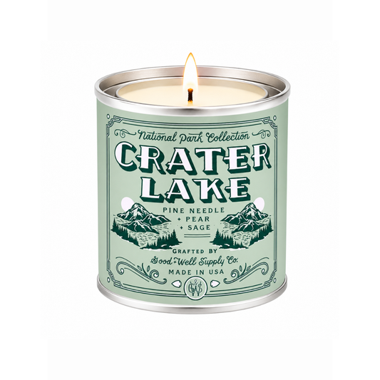 A lit scented candle in a cylindrical metal tin with a pale sage green label reading 'National Park Collection Crater Lake' and mountain illustrations, on a white background.