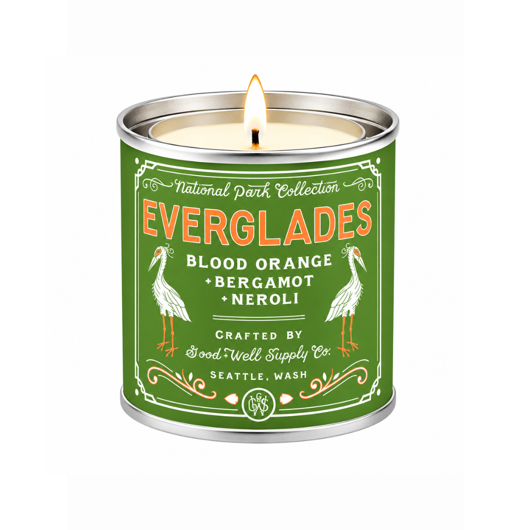 A lit candle in a cylindrical metal tin with a green label reading 'Everglades Blood Orange Bergamot Neroli' and illustrated birds, on a white background.