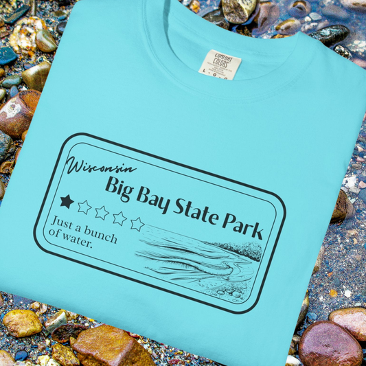 A folded Lagoon Blue Colored T-Shirt with 1 star review design of Big Bay State Park.  Features a line art image of a sandy beach and shoreline, 1 star graphic, and the text: “Wisconsin Big Bay State Park, Just a bunch of water.  T-shirt is folded and sitting on rounded lake pebbles.