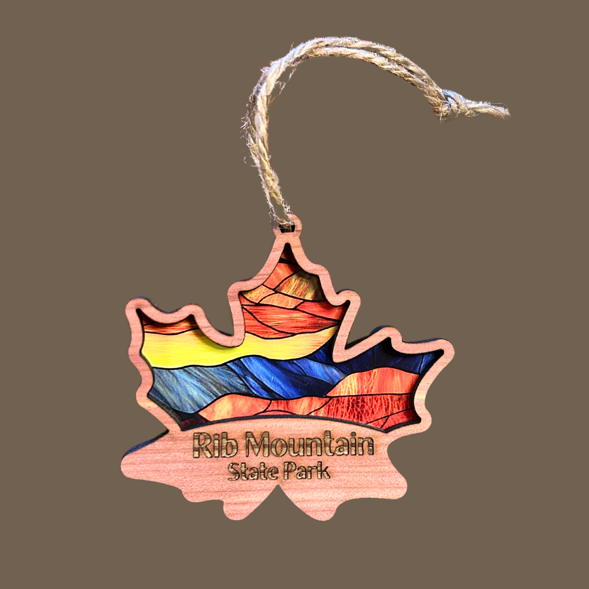 Cedar wood maple leaf ornament with stained glass appearance and the text “Rib Mountain State Park.”  Photo Background: brown.