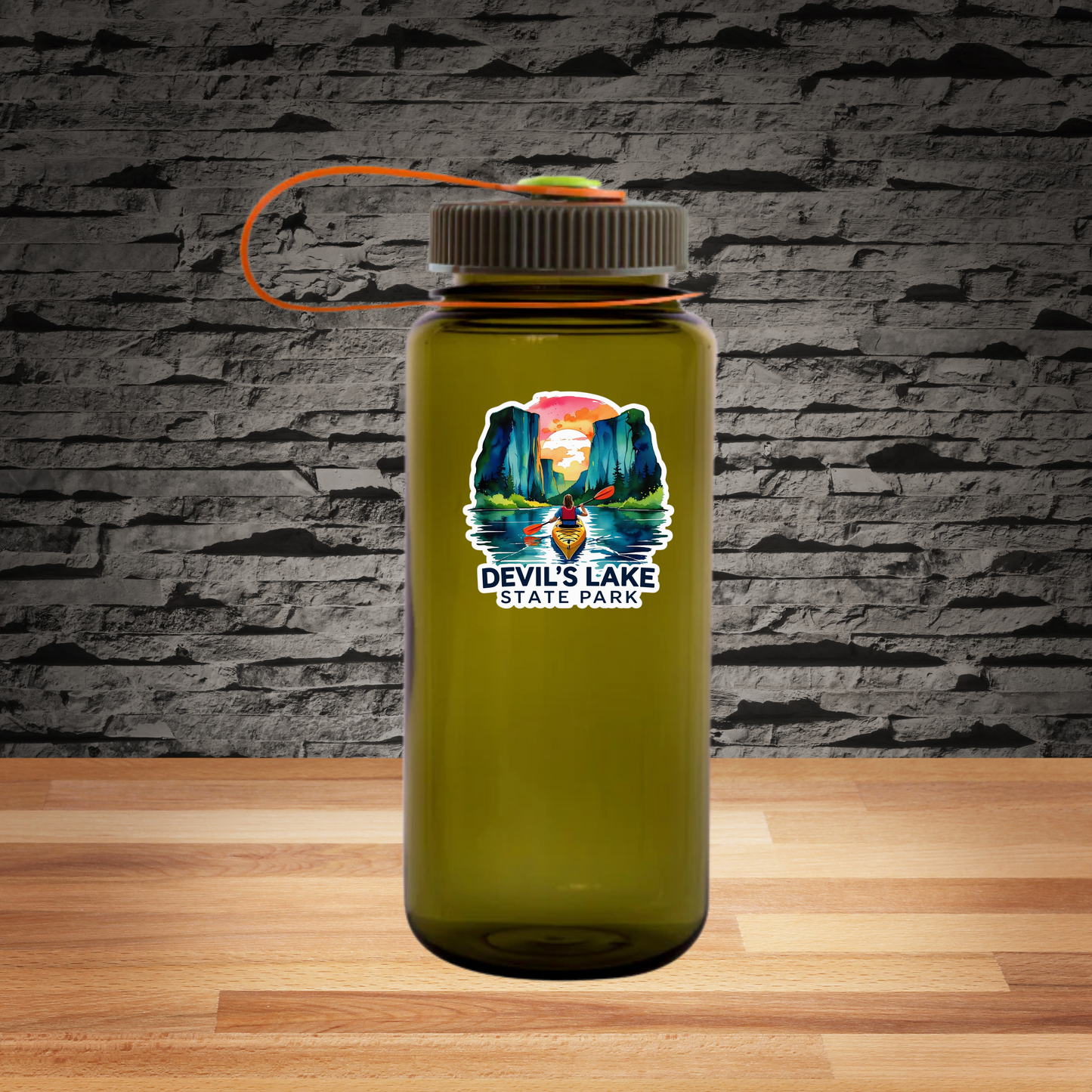 A water bottle sitting on a wood table top in front of a rock wall with a Watercolor style sticker featuring a kayaker on a lake surrounded by towering rock bluffs, a vivid sunset and the text Devil’s Lake State Park. 