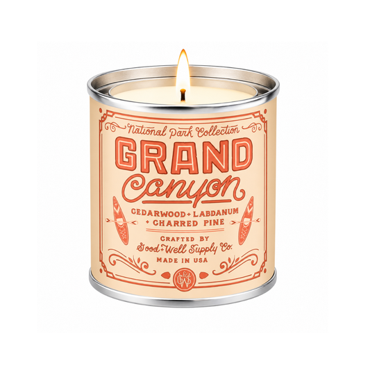 A lit scented candle in a cylindrical metal tin with a cream-colored label featuring red-orange text that reads 'GRAND Canyon' and smaller scent notes including Cedarwood, Labdanum, and Charred Pine. The candle is photographed on a clean white background.