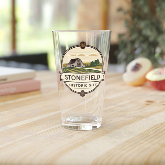16oz pint glass featuring a logo design of a barn set amongst fields.  Photo background: glass sitting in a wood counter with books and fruit.