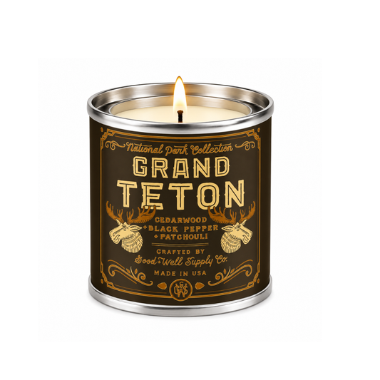 A lit scented candle in a cylindrical metal tin with a dark brown label featuring gold text and antler illustrations. The label reads 'National Park Collection GRAND TETON' and 'CEDARWOOD BLACK PEPPER PATCHOUli'.