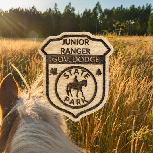 Laser engraved wood badge featuring the text: “Junior Ranger Gov Dodge State Park” with the icon of a person riding a horse.  Photo Background: Horse walking through a golden field of tall grass.