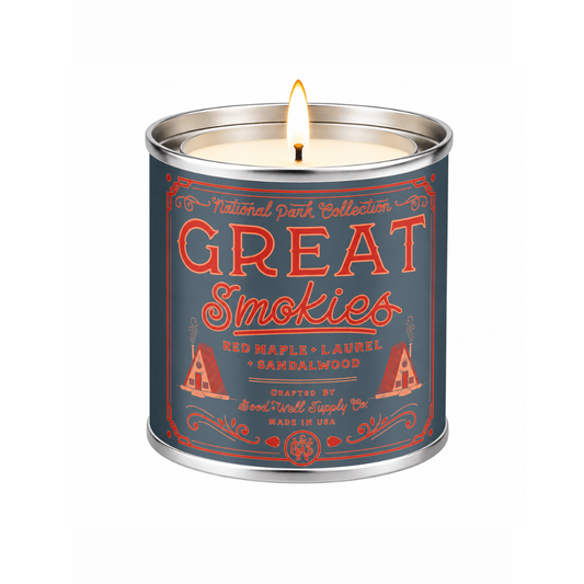 A lit scented candle in a cylindrical metal tin with a dark blue label and red-orange text reading “Great Smokies,” shown on a plain white background.