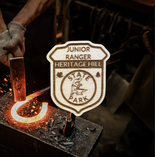 Wood badge shaped JR Ranger Badge with the text “Junior Ranger, Heritage Hill State Park” with a blacksmith symbol.  Photo background: blacksmith forging a hot iron hook.