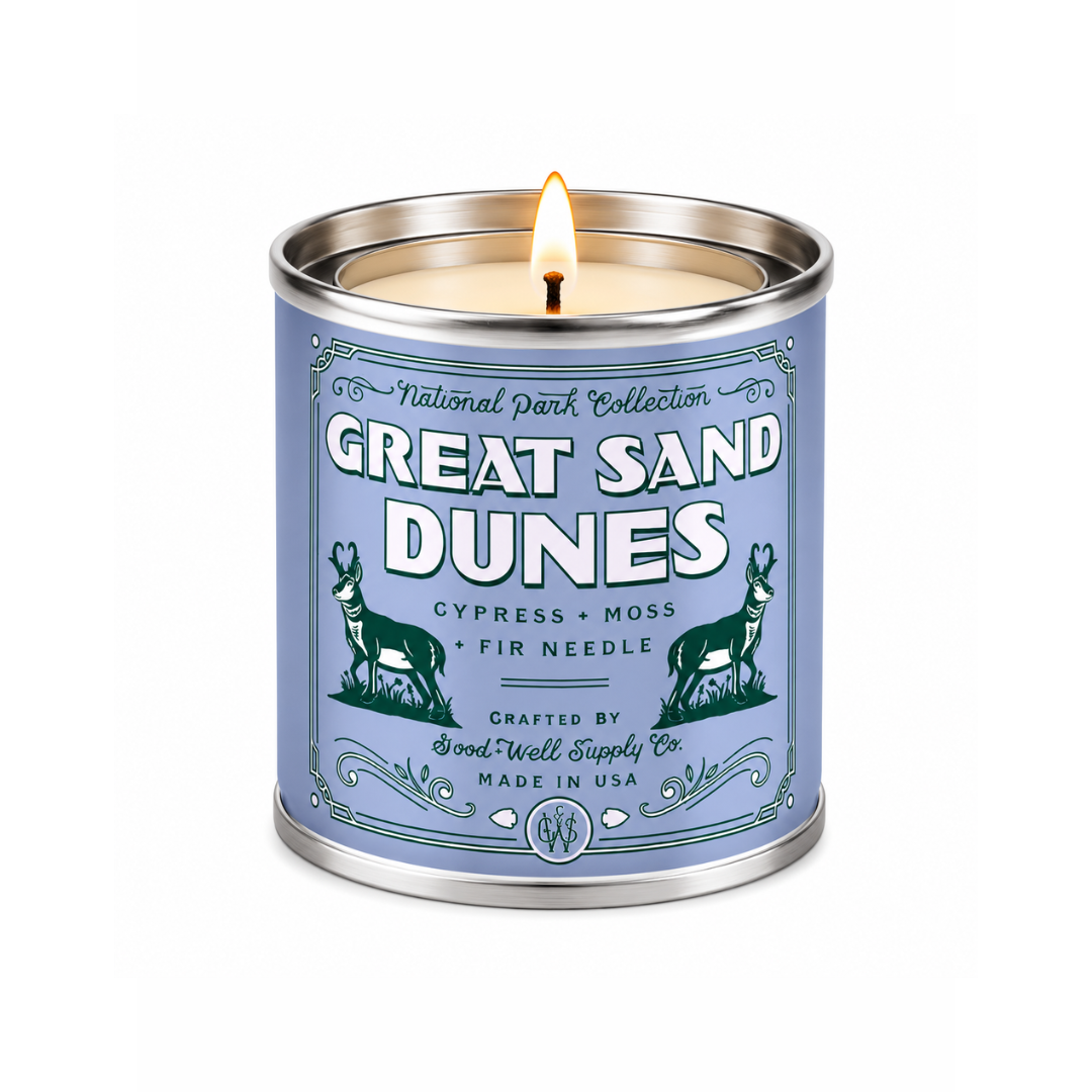 A lit scented candle in a cylindrical metal tin with a light blue label reading 'Great Sand Dunes' and 'National Park Collection.' The tin has a silver metal rim and base, and the candle is shown on a plain white background.