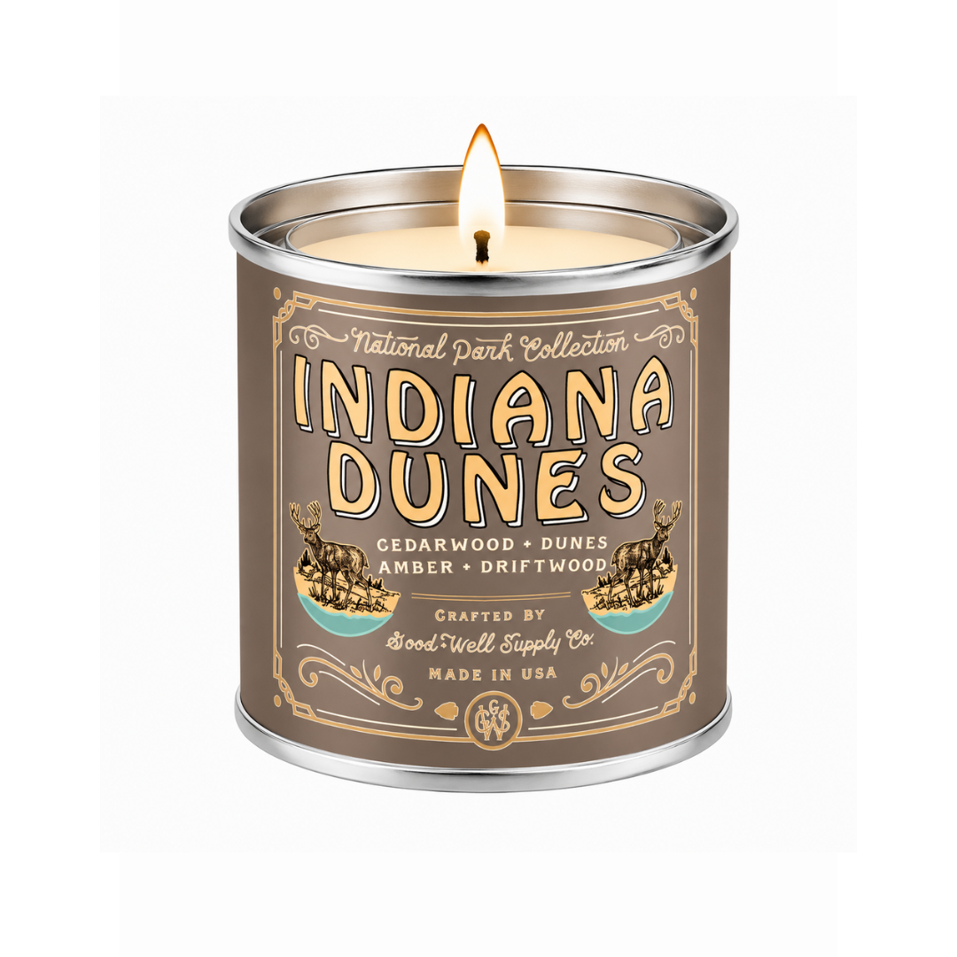 A lit scented candle in a cylindrical metal tin with a brown label reading 'Indiana Dunes' and 'National Park Collection.' The candle is shown on a clean white background, with the flame visible above the rim.