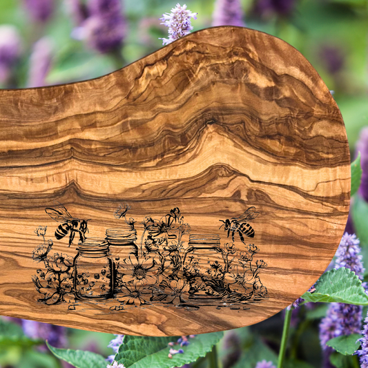Natural olive wood serving board with a laser engraved scene depicting bees, flowers, and mason jars.  Photo background:  Purple flowers.