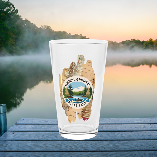 16 oz glass pint glass featuring a birch bark design with a depiction Native Americans in a birch bark canoe paddling on a river.  Photo Background: glass sitting at the end of a dock on foggy morning.