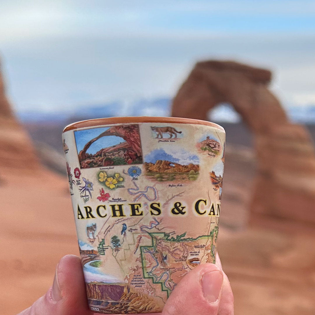 A hand holds a small illustrated shot glass featuring desert landscapes and the text 'Arches & Canyonlands’ in front of a blurred background of Delicate Arch and red rock formations.