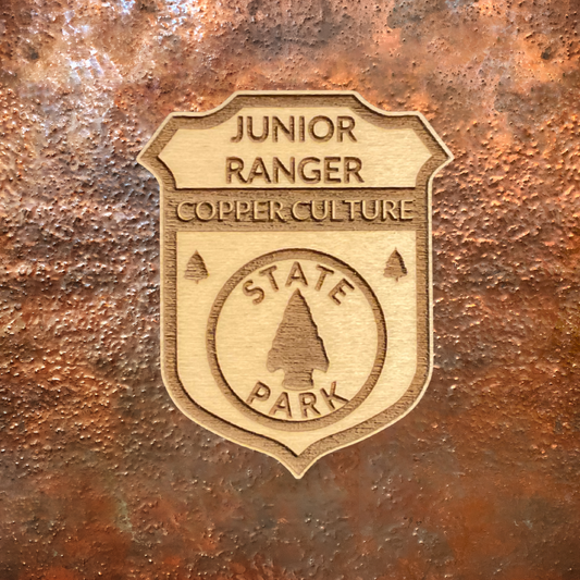 Wood Shaped badge with the text “Junior Ranger Copper Culture State Park” and an arrowhead symbol.  Photo background: copper metal.
