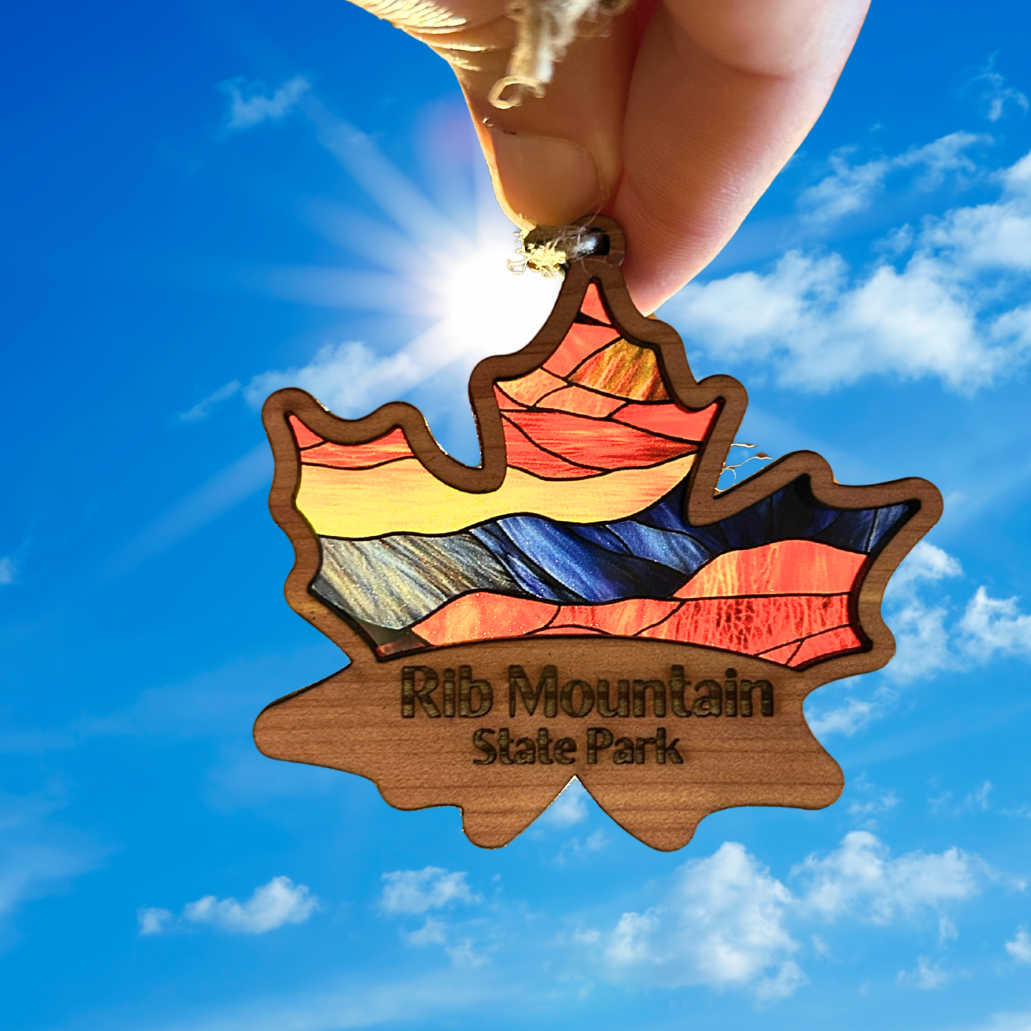 Cedar wood maple leaf ornament with stained glass appearance and the text “Rib Mountain State Park.”  Photo Background: ornament being held up to the sun to show the vibrant design.