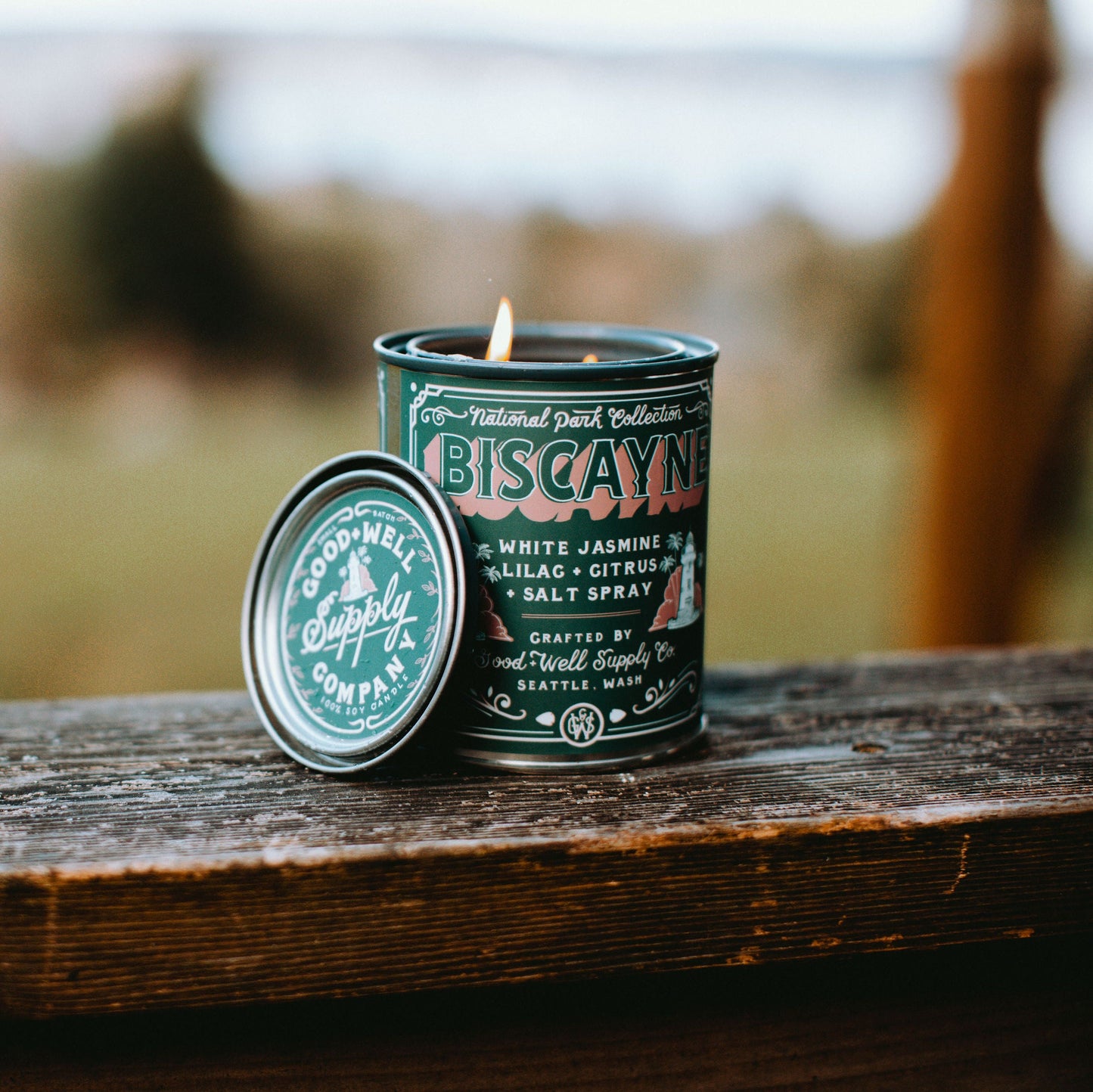 Candle in a decorative tin labeled 'Biscayne' on a wooden surface with a blurred natural background