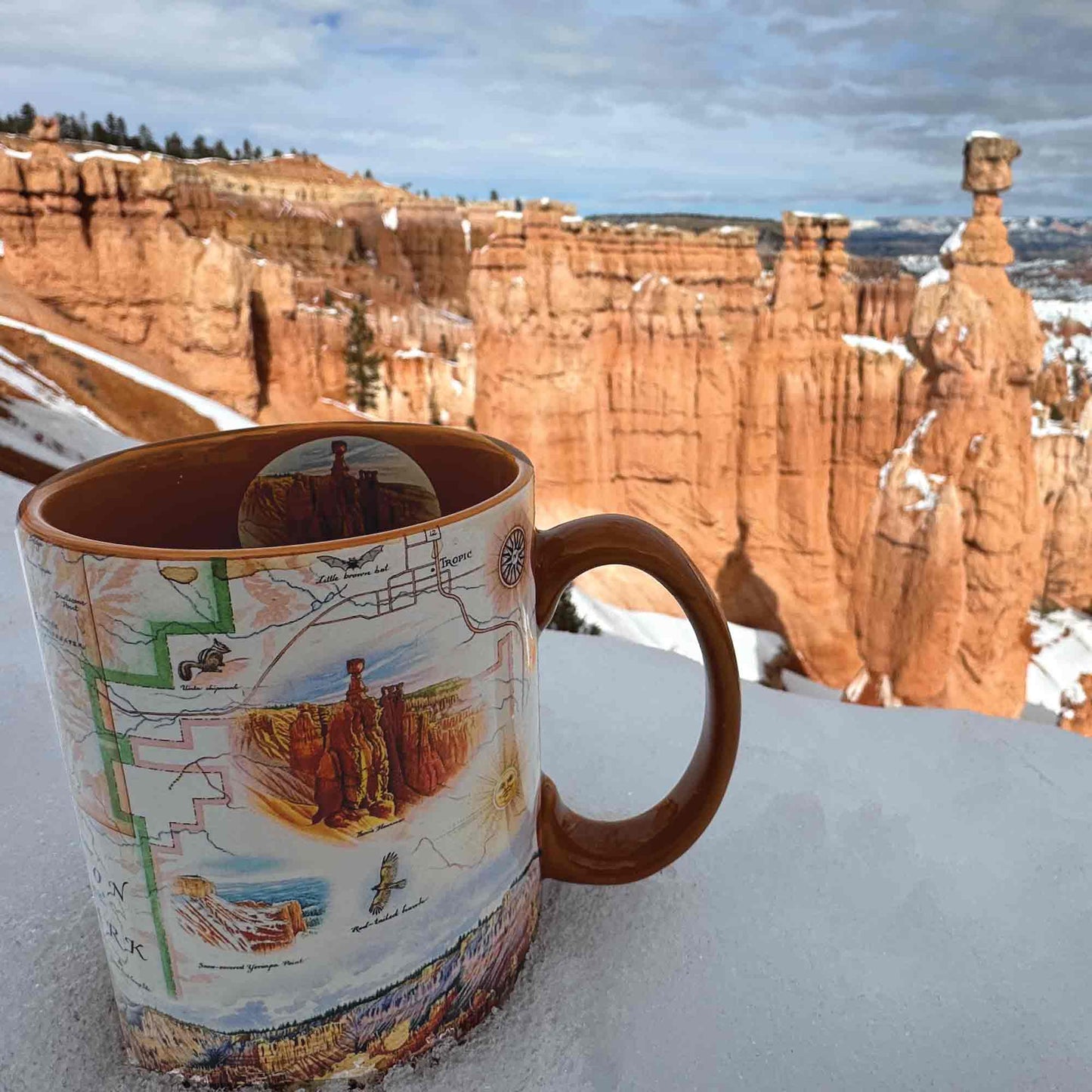 A ceramic mug with a detailed map and landscape illustration, featuring a brown handle and interior, set against a snowy outdoor background of hoodoos and rock formations.