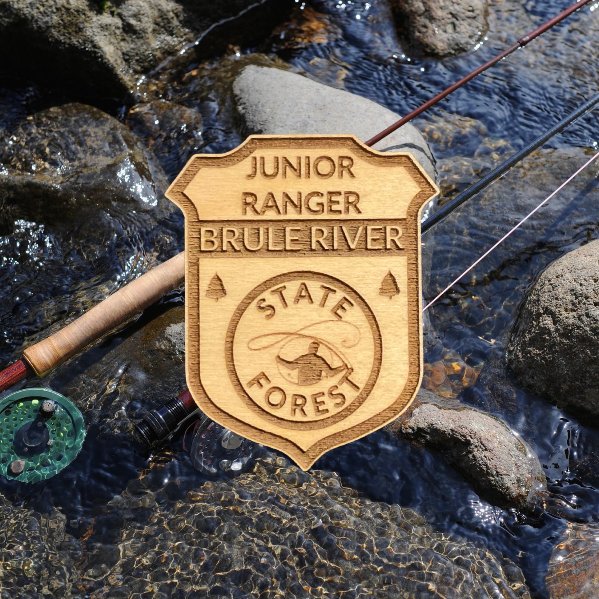 Wood Jr Ranger badge with the text: “Junior Ranger Brule River State Forest” and an icon of a fly fisherman casting.  Photo background: two fly rods laying on the rocks in a stream.