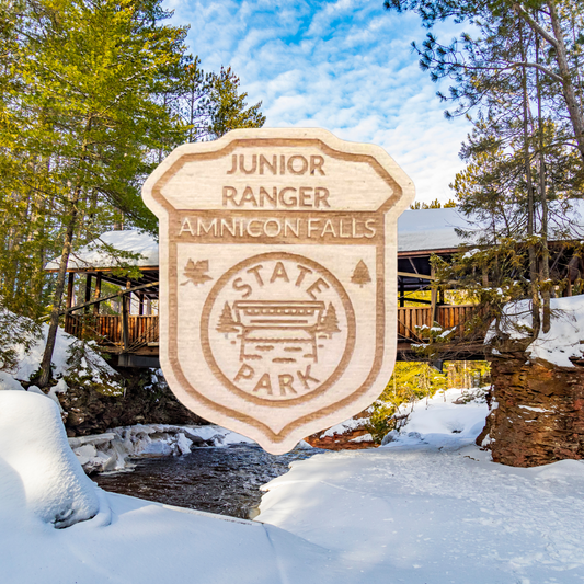 Wood shaped Jr. Ranger Badge depicting a covered bridge over a stream and the text: Junior Ranger Amnicon Falls State Park.  Photo Background: covered bridge over a partially frozen river.