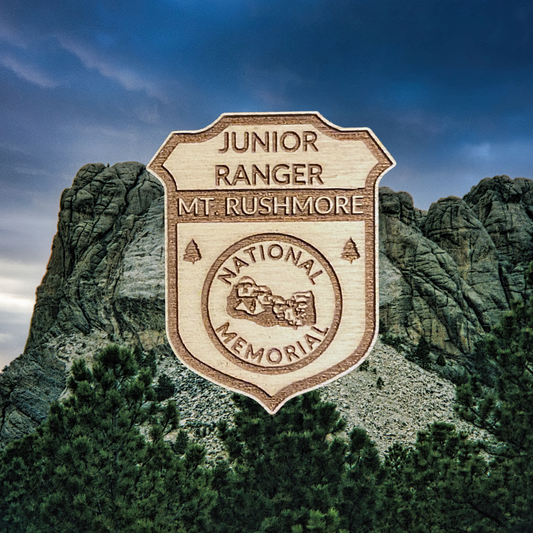 Wood Junior Ranger badge featuring an icon of Mount Rushmore with the Text: Junior Ranger Mount Rushmore National Memorial.  Photo background: Mount Rushmore against a dark cloudy sky.