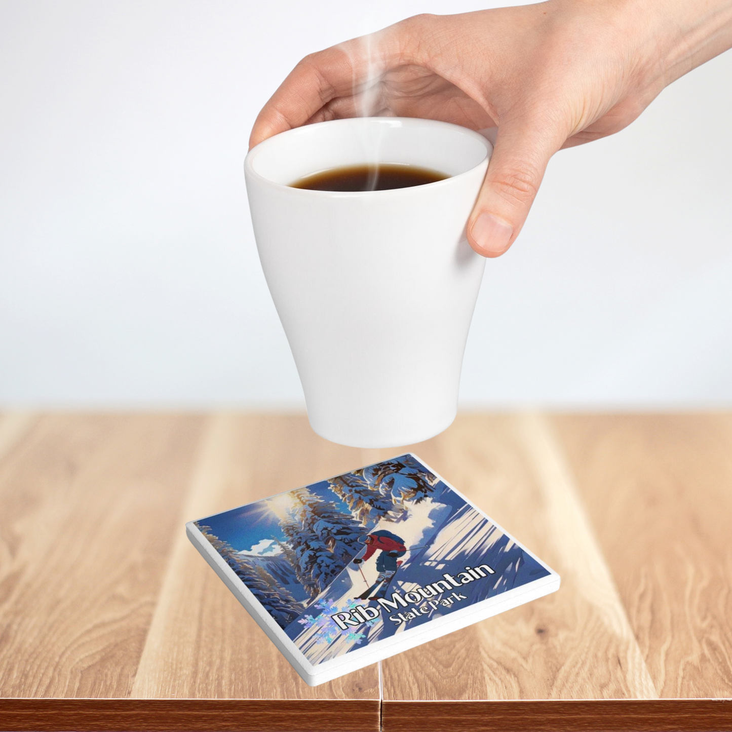 Ceramic coaster featuring a skier going down a snowy, wooded slope and the text: Rib Mountain State Park.  Photo background: person setting a steaming hot beverage on a coaster. 