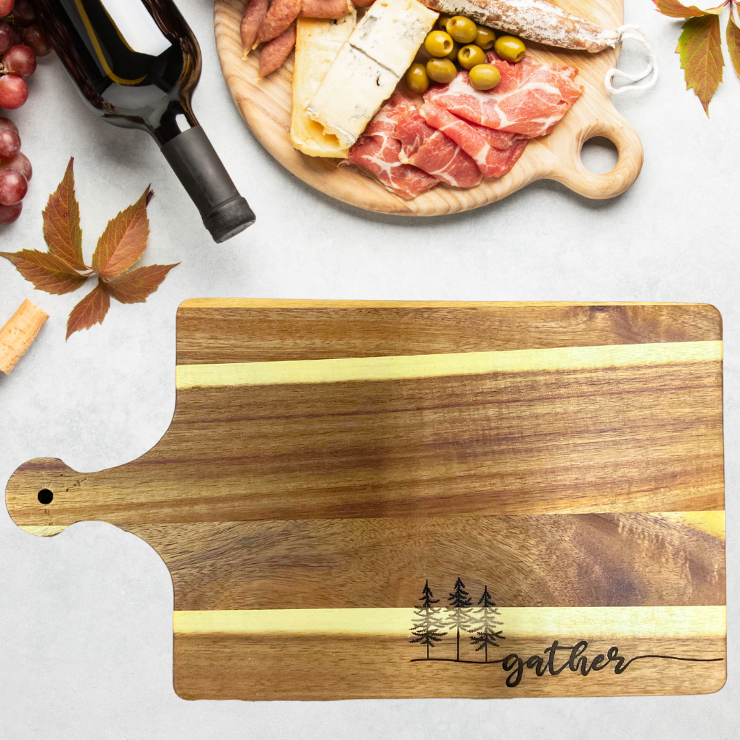 Acacia wood cutting board with three trees and the text gather engraved on the board.  Wine and cheese background. 