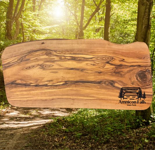 Olive wood serving board with a graphic depicting a covered bridge over a stream with trees and the text Amnicon Falls State Park.  Photo background: hiking trail through a lush green forest.