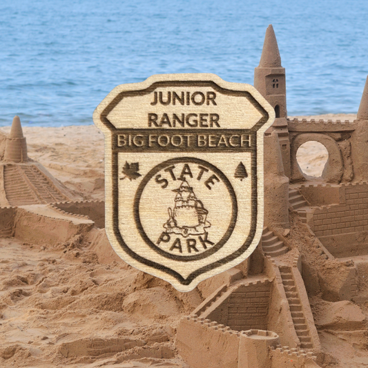 Laser engraved wood badge featuring the text: “Junior Big Foot Beach State Park” with the icon of a sandcastle.  Photo Background: sandcastle built on a sandy beach next to the water.