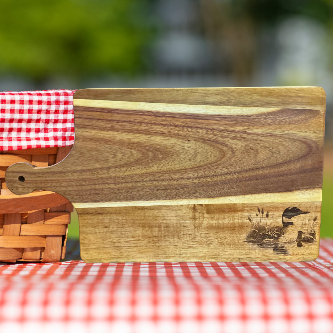 Acacia wood cutting board with a loon and two chicks engraved on it.  Picnic table and picnic basket background. 