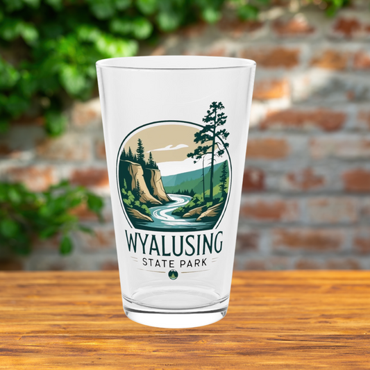 Glass pint glass featuring a design of a winding river below a bluff.  Photo Background: glass sitting on a wood table in front of a brick wall. 