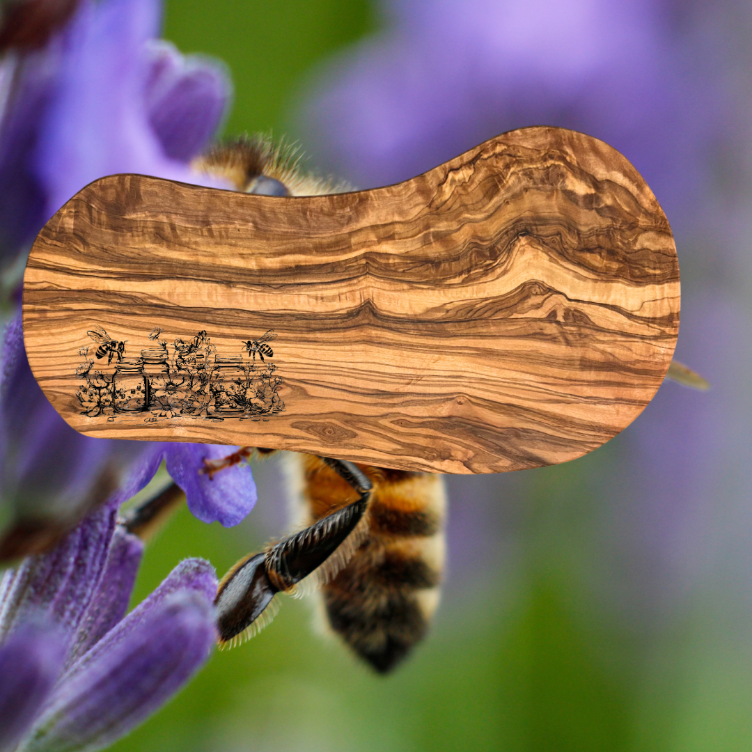 Natural olive wood serving board with a laser engraved scene depicting bees, flowers, and mason jars.  Photo background:  Purple flowers and a bee.