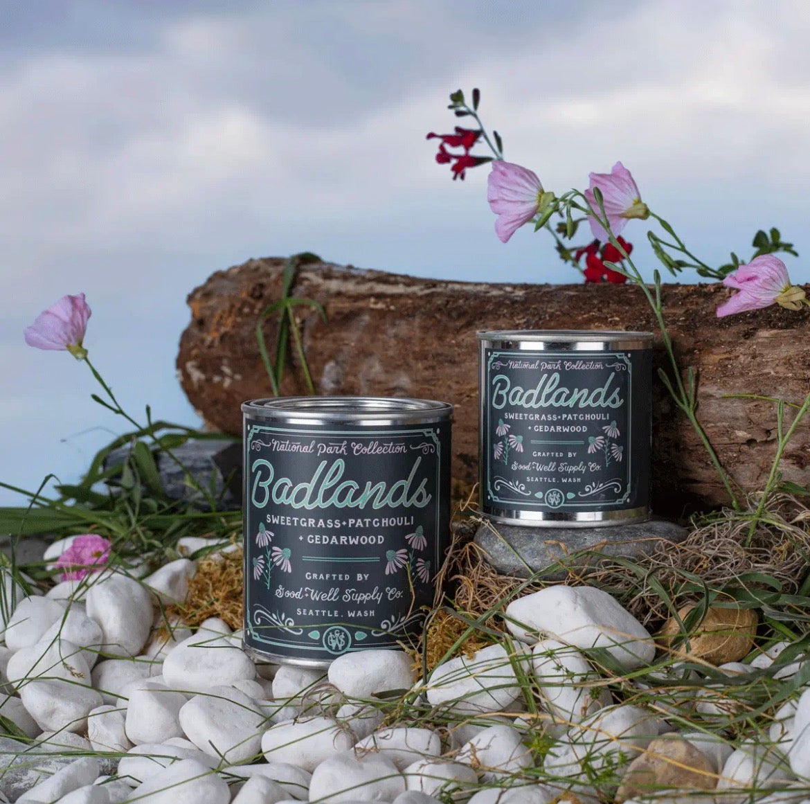 Two Badlands Candle tin cans displayed outdoors on white stones with a log and pink flowers in the background. The cans are dark gray with white script lettering and are labeled Sweetgrass & Cedarwood and Sweetgrass & Patchouli.