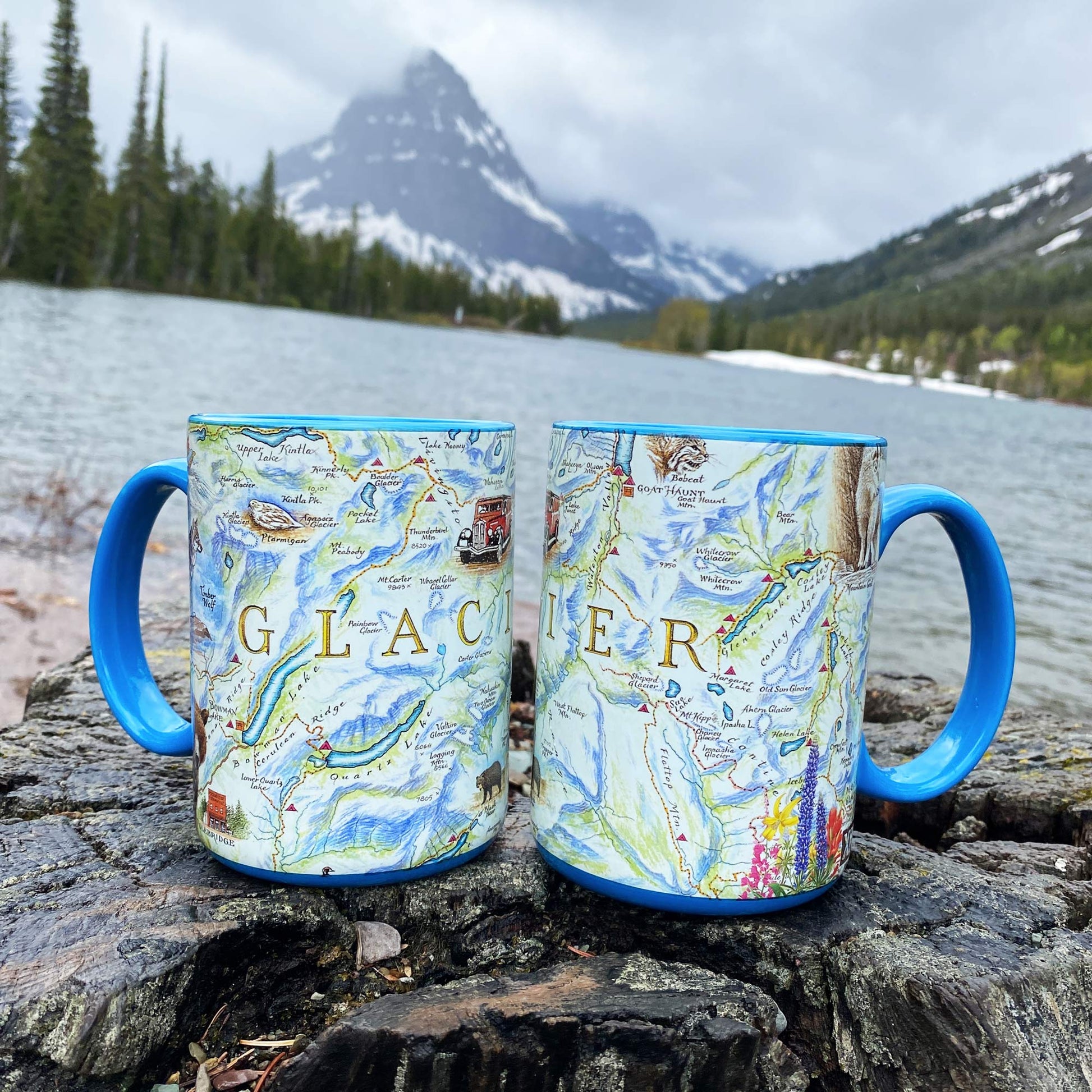 Two ceramic mugs with blue handles and blue rims sit on a rock by a lake, printed with a scenic Glacier map design and the word "GLACIER" across the front, with a mountain and forest in the background.