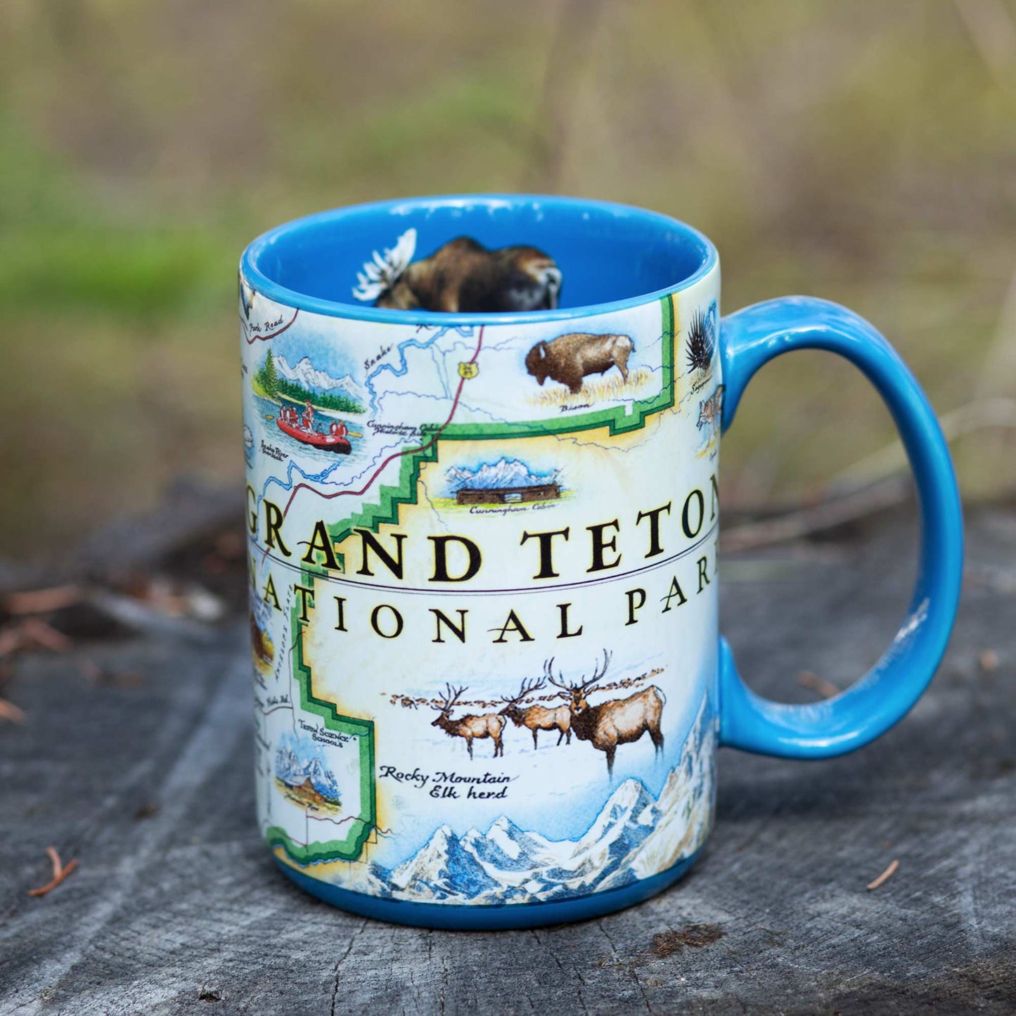 A ceramic mug featuring a colorful illustrated map of Grand Teton National Park with bison, elk, mountains, and the park name, set on a weathered wooden surface with a blurred outdoor background.