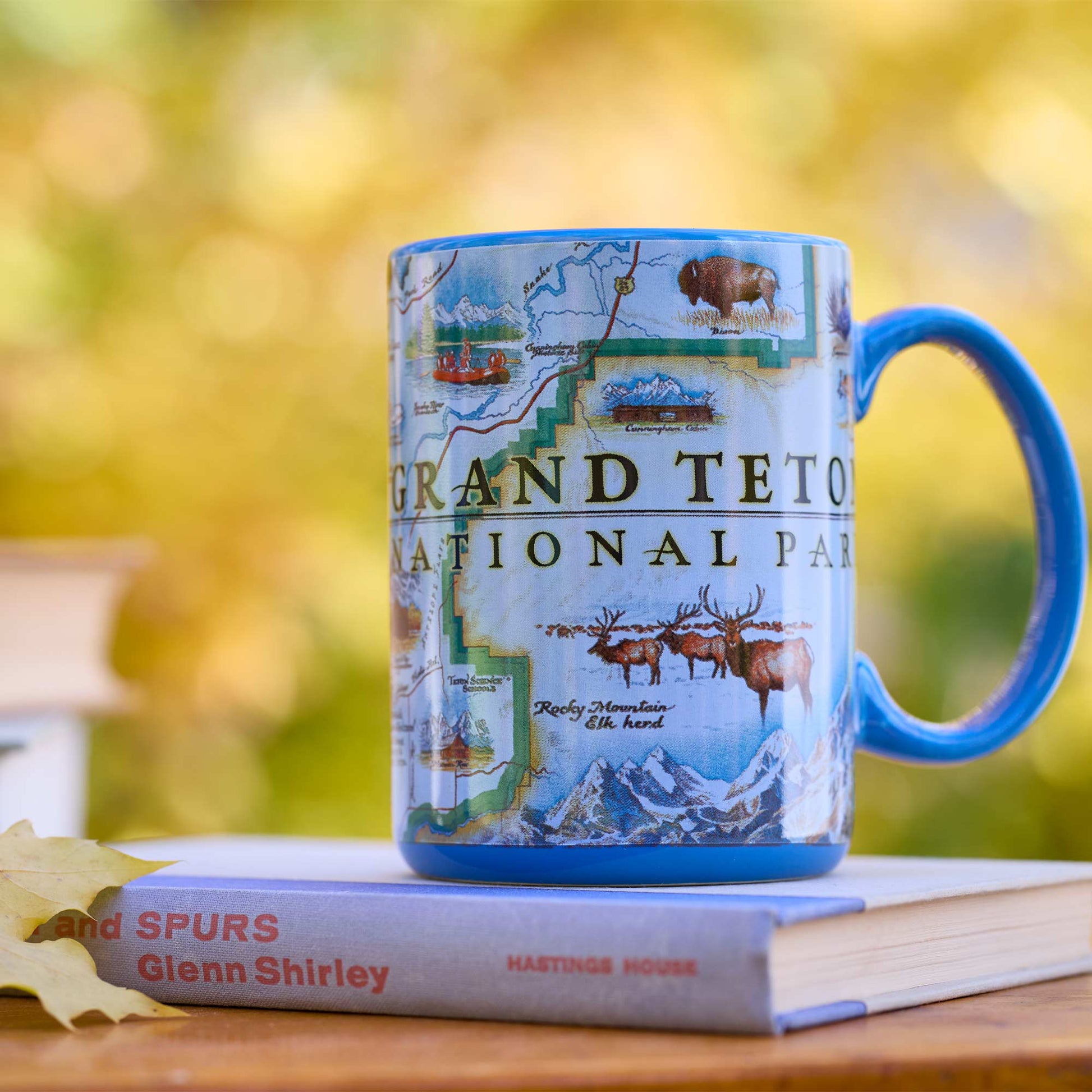 A blue ceramic mug printed with a Grand Teton National Park map design, resting on top of a book on a wooden surface with a blurred outdoor background.