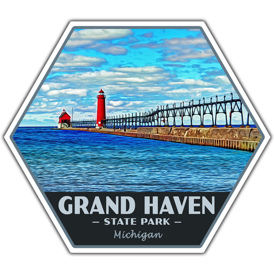 A hexagonal sticker or decal featuring a stylized illustration of Grand Haven State Park in Michigan, with a red lighthouse, pier, blue sky, and the text 'GRAND HAVEN STATE PARK Michigan' on a dark band at the bottom.