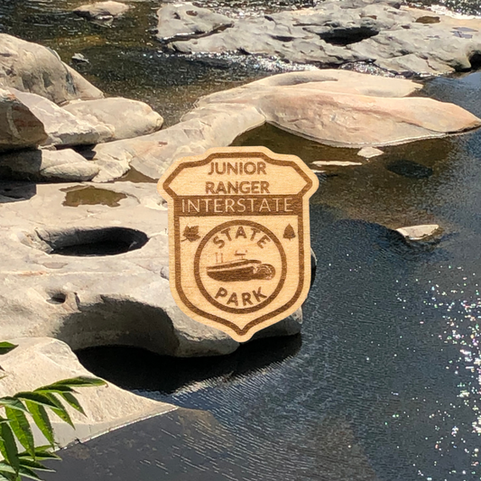 Wood badge shaped pin featuring the text “Junior Ranger - Interstate State Park” with an image of a paddle wheel boat.  Photo background: pothole rock formations along a river.