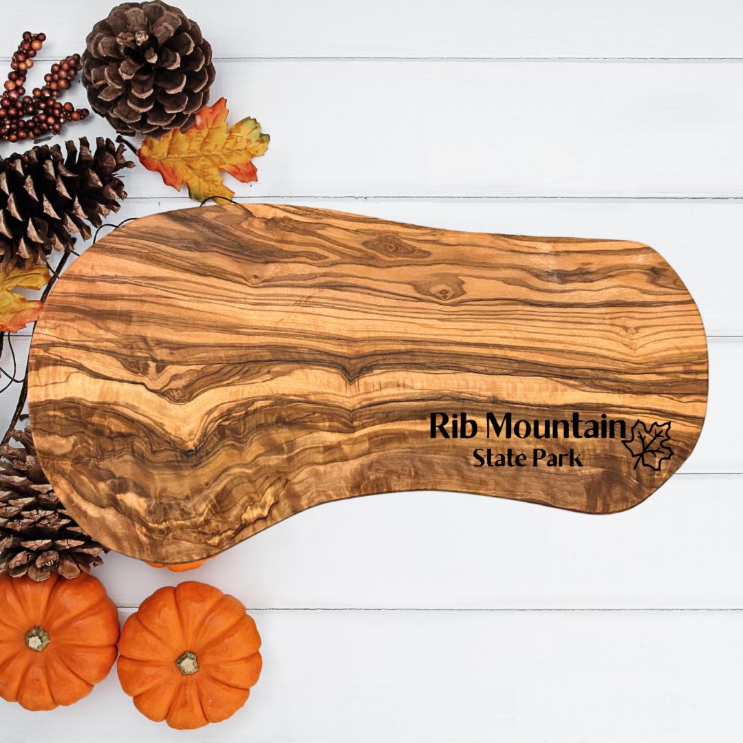 Natural olive wood serving board with the text “Rib Mountain State Park” and a maple leaf symbol engraved on it.  Photo background: white shiplap with leaves, pine cones and miniature pumpkins..