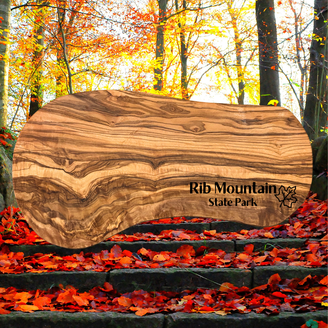 Natural olive wood serving board with the text “Rib Mountain State Park” and a maple leaf symbol engraved on it.  Photo background: vibrant red leaves lying on stone steps leading into the woods.