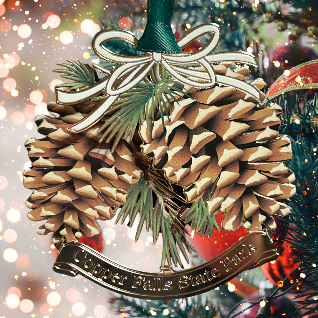 Handcrafted brass ornament featuring two pinecones and pine branches.  Features a custom banner with the text Copper Falls State Park. Photo background: Christmas tree and lights.