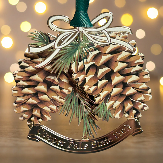 Handcrafted brass ornament featuring two pinecones and pine branches.  Features a custom banner with the text Copper Falls State Park. Photo background: white Christmas lights.