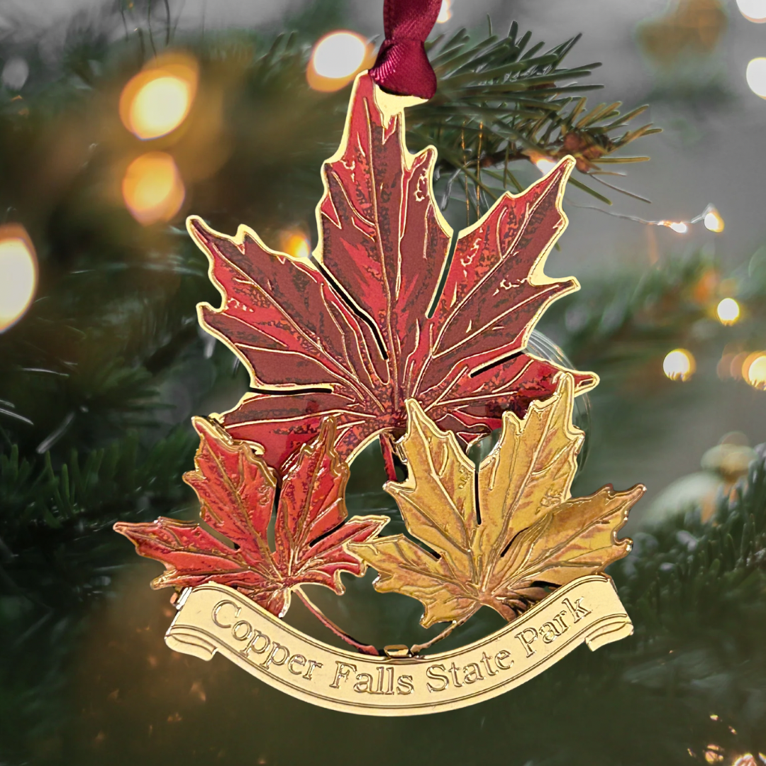 Handcrafted brass ornament featuring three vibrant maple leafs in red, orange, and yellow. Features a custom banner with the text Copper Falls State Park.  Photo background: Christmas tree and lights.