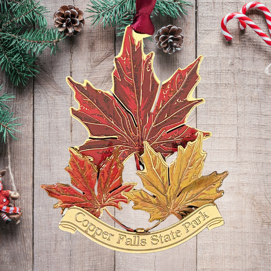 Handcrafted brass ornament featuring three vibrant maple leafs in red, orange, and yellow. Features a custom banner with the text Copper Falls State Park.  Photo background: wood boards and garland.