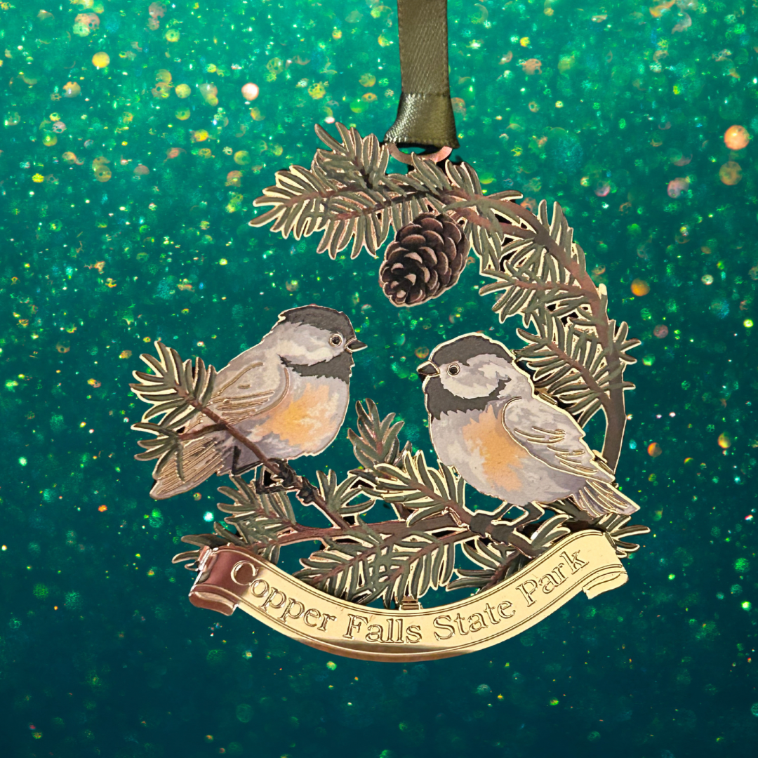 Handcrafted brass ornament featuring two chickadees sitting on pine needle branches.  Features a custom banner with the text Copper Falls State Park. Photo background: green glitter.