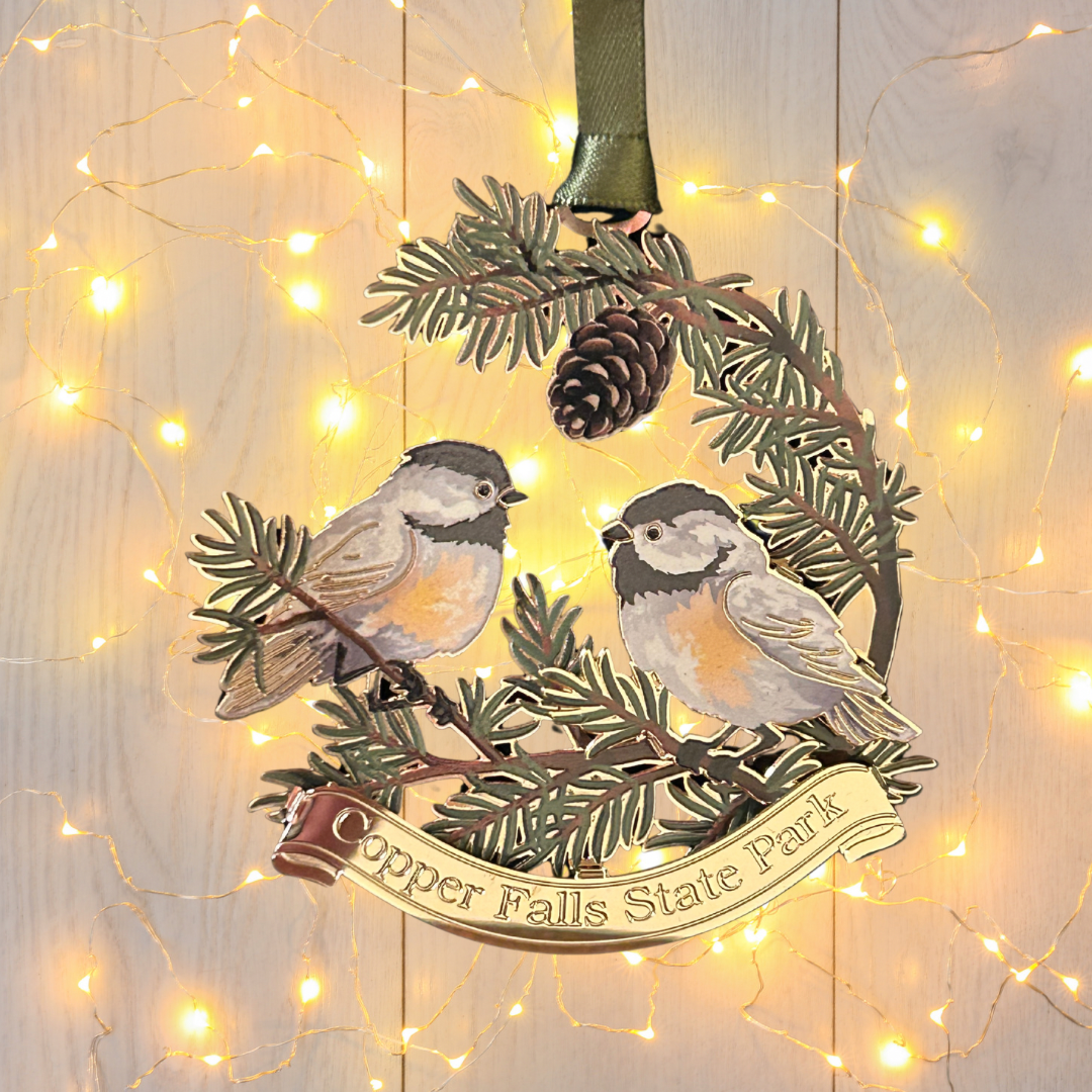 Handcrafted brass ornament featuring two chickadees sitting on pine needle branches.  Features a custom banner with the text Copper Falls State Park. Photo background: white washed wood and white lights.