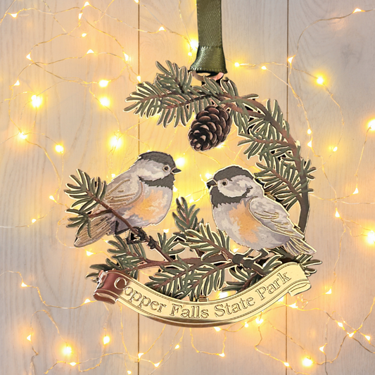 Handcrafted brass ornament featuring two chickadees sitting on pine needle branches.  Features a custom banner with the text Copper Falls State Park. Photo background: white washed wood and white lights.