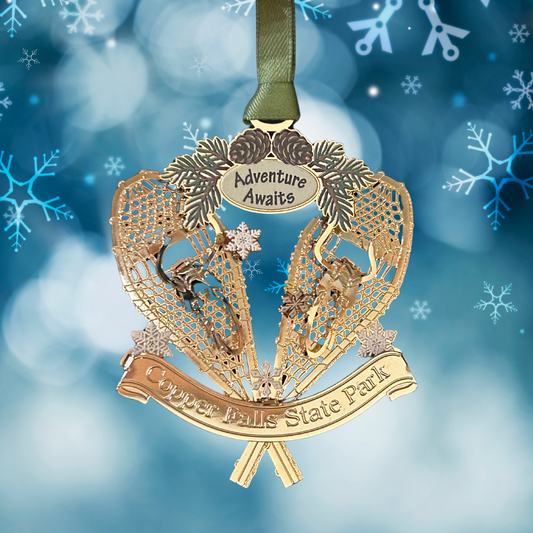 Handcrafted brass ornament featuring two crossed snowshoes with pine cones and boughs.  Features a custom banner with the text Copper Falls State Park and adventure awaits.  Photo background: blue background and snowflakes.