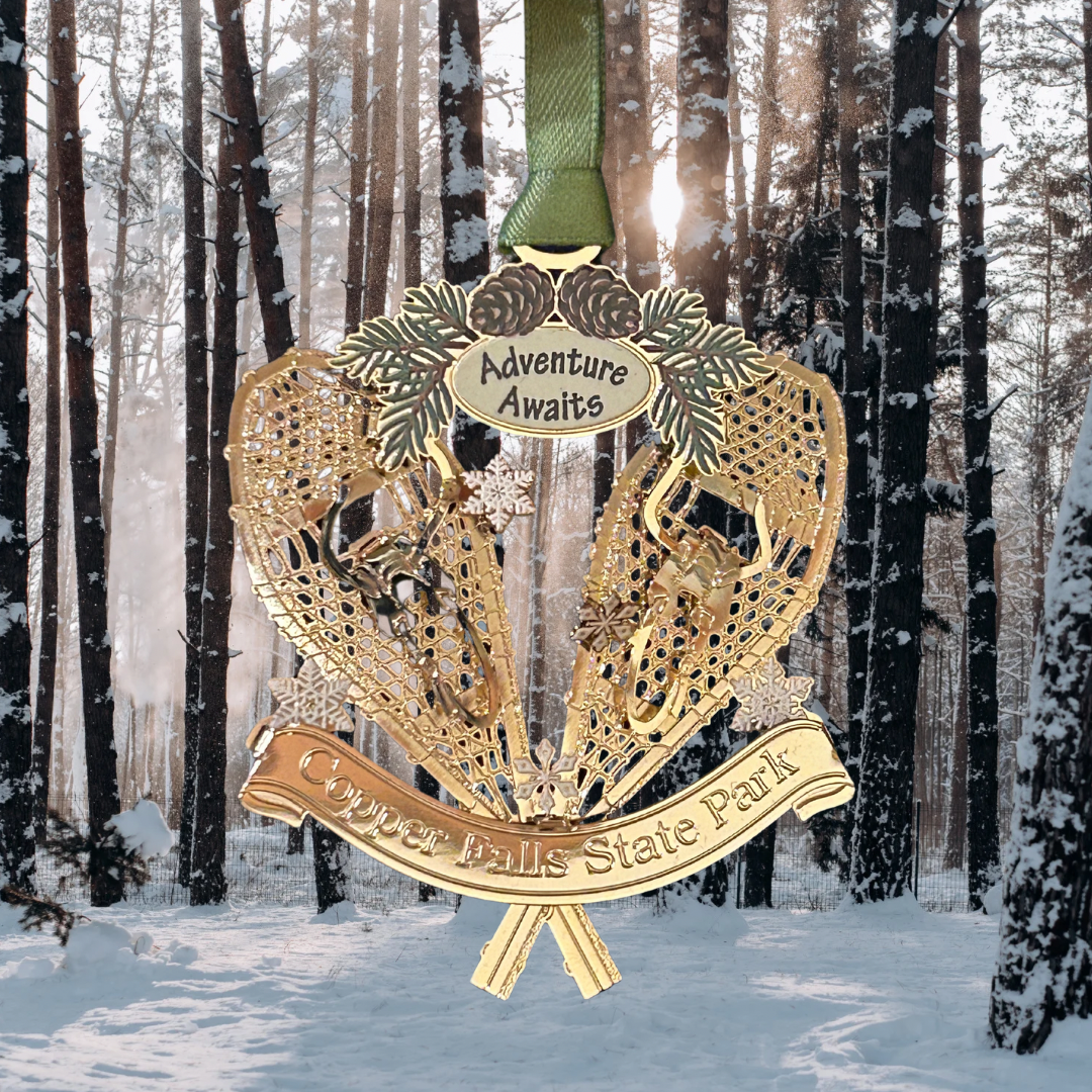 Handcrafted brass ornament featuring two crossed snowshoes with pine cones and boughs.  Features a custom banner with the text Copper Falls State Park and adventure awaits.  Photo background: snowy woods.